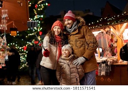 family, winter holidays and technology concept - happy mother, father and little daughter taking picture by smartphone on selfie stick at christmas market on town hall square in tallinn, estonia