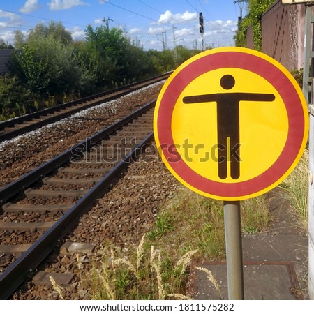 YRound yellow sign or signal with black human figure with outstretched arms and red borders marking end of train platform at railroad station in Germany. Rail tracks with traffic lights in background