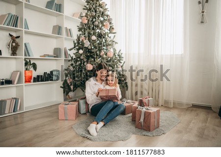 Beautiful mother and daughter open Christmas presents at home near the Christmas tree in a white interior. Family happiness, holiday, joy, vacation.