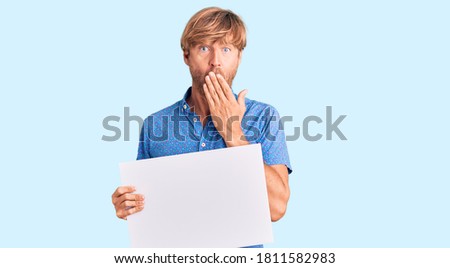 Handsome caucasian man with beard holding sale poster covering mouth with hand, shocked and afraid for mistake. surprised expression 