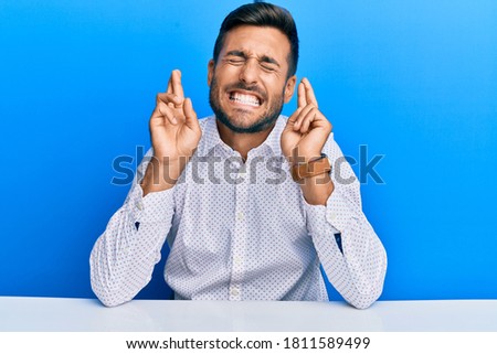 Handsome hispanic man wearing business clothes sitting on the table gesturing finger crossed smiling with hope and eyes closed. luck and superstitious concept. 