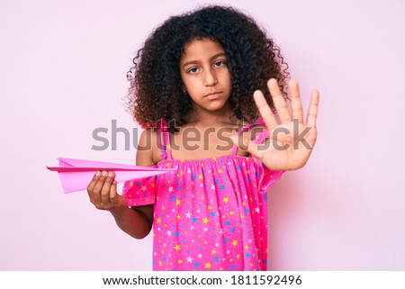 African american child with curly hair holding paper airplane with open hand doing stop sign with serious and confident expression, defense gesture 