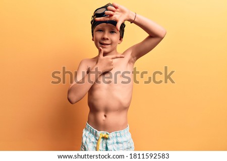 Cute blond kid wearing swimwear and swimmer glasses smiling making frame with hands and fingers with happy face. creativity and photography concept. 