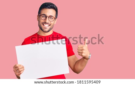Young hispanic man holding blank empty banner smiling happy and positive, thumb up doing excellent and approval sign 