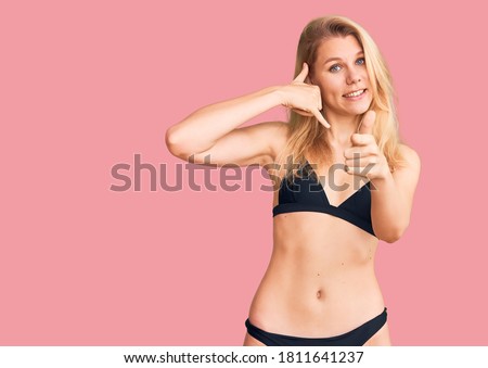 Young beautiful blonde woman wearing bikini smiling doing talking on the telephone gesture and pointing to you. call me. 