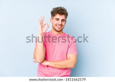 Young blond curly hair caucasian man isolated winks an eye and holds an okay gesture with hand.