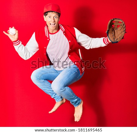 Young handsome man wearing sporty clothes smiling happy. Jumping with smile on face playing baseball using ball and glove over isolated red background