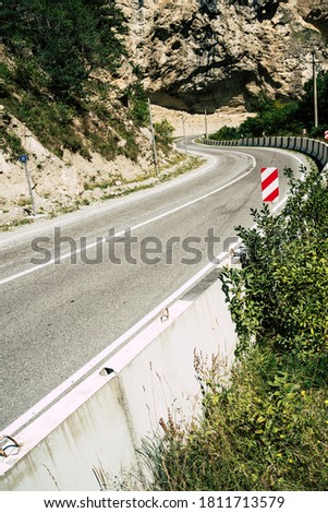 Curvy road with traffic signs in mountains. Red diagonal keep left obstacle traffic sign placed on stone border of curvy asphalt road going through mountainous terrain