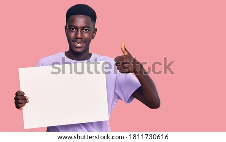 Young african american man holding blank empty banner smiling happy and positive, thumb up doing excellent and approval sign 