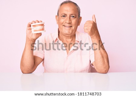 Senior handsome man with gray hair holding denture sitting on the talbe smiling happy and positive, thumb up doing excellent and approval sign 