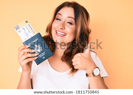 Young beautiful caucasian woman holding boarding pass and passport smiling happy and positive, thumb up doing excellent and approval sign 