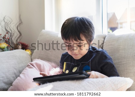High key kid sitting on sofa watching cartoon on tablet,Happy boy playing game on touch pad with bright light in morning. ute Kid having fun and relaxing on his own in living room,New normal lifestyle