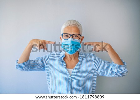Mature woman wearing a face mask on gray background. Flu epidemic and virus protection concept. Safety during a pandemic, epidemic, seasonal flu. Woman points to medical mask on her face