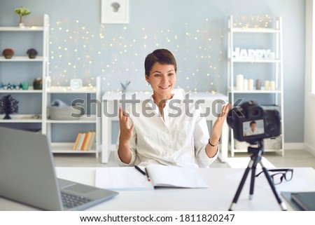 Positive female vlogger recording a video on camera sitting at desk in modern office. Happy businesswoman sharing experience making a pre-recorded webinar at home