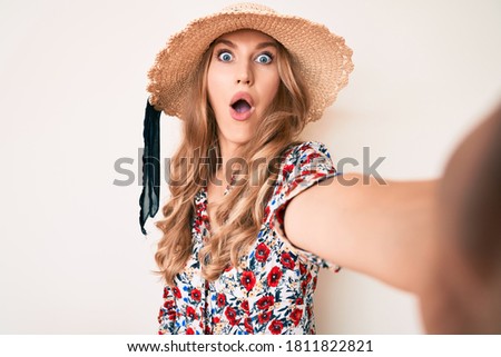 Young caucasian woman with blond hair wearing summer hat taking a selfie scared and amazed with open mouth for surprise, disbelief face 