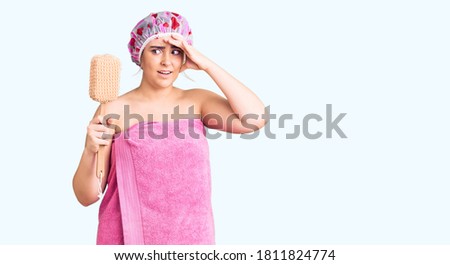 Young caucasian woman wearing shower cap and towel holding sponge stressed and frustrated with hand on head, surprised and angry face 