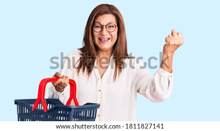 Middle age latin woman holding supermarket shopping basket screaming proud, celebrating victory and success very excited with raised arms 