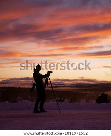 A silhouette of a young photographer shooting a snowy landscape at sunset with a camera on a tripod