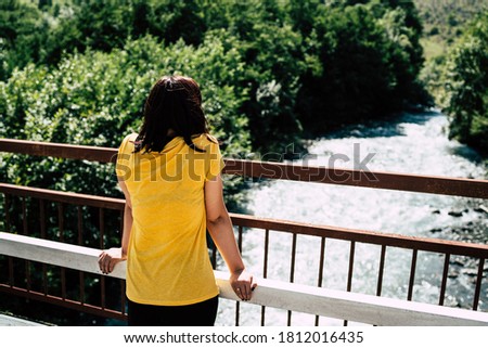 Unrecognizable young woman stands on bridge and enjoys beautiful view of mountain river which flowing between green trees. Rear view of female tourist looking at clean waterway