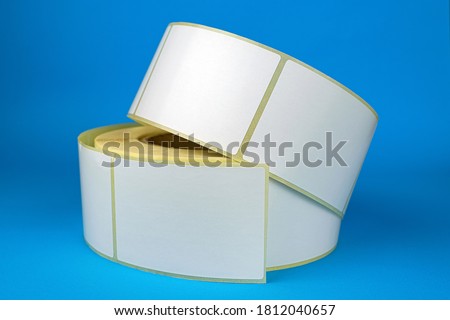 Babina of self-adhesive stickers on a blue background. White roll of labels for thermal perforation Self-adhesive white label roller for printing or manufacturing Royalty-Free Stock Photo #1812040657