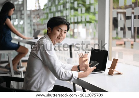 Business man smiling and holding smart phone while browsing some interesting content on  laptop. He is sitting in a coffee shop.