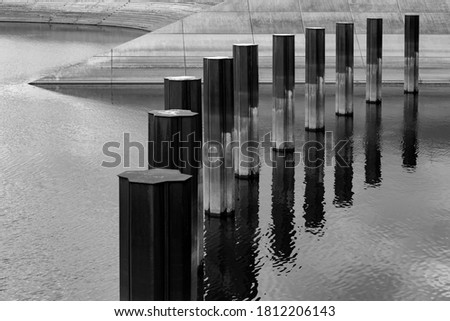 Bollards or Capstans in a bent row and concrete Bank fortifications in the background in the inland port of Duisburg with reflection in the water surface black and white 
