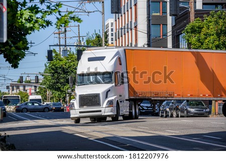 White classic industrial diesel big rig semi truck tractor transporting commercial cargo in orange dry van semi trailer turning on the crossroad street intersection with busy city traffic Royalty-Free Stock Photo #1812207976