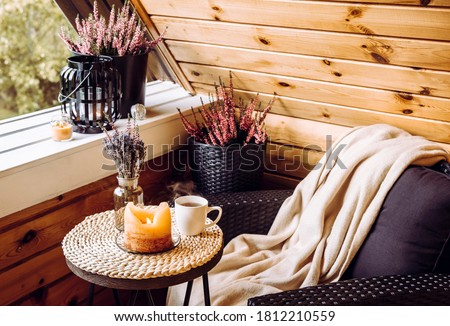 Cute autumn hygge home decor arrangement. Tiny wooden cabin balcony with heather flowers, lavender in bottle vase, candlelight flame, soft beige plaid waiting on comfortable garden furniture chair. Royalty-Free Stock Photo #1812210559