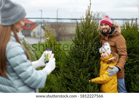 family, winter holidays and people concept - happy mother, father and little daughter buying christmas tree and taking picture with smartphone at street market