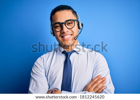 Young brazilian call center agent man wearing glasses and tie working using headset happy face smiling with crossed arms looking at the camera. Positive person. Royalty-Free Stock Photo #1812245620