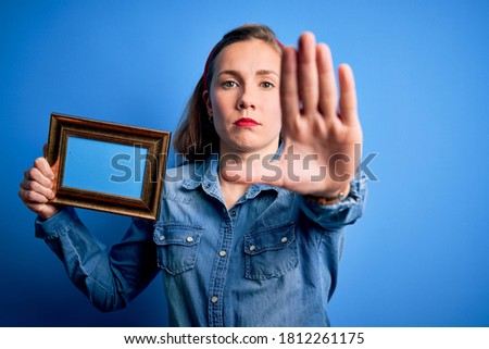 Young beautiful blonde woman holding vintage frame over isolated blue background with open hand doing stop sign with serious and confident expression, defense gesture
