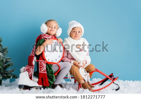 kids in winter outfit and scarfs sitting on sleigh near clock, pine and ice skates on blue