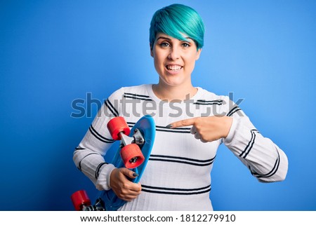 Young skater woman with blue fashion hair holding skateboard over blue isolated background very happy pointing with hand and finger