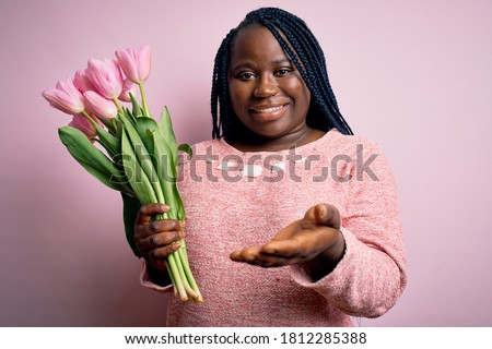 Young african american plus size woman with braids holding bouquet of pink tulips flower smiling cheerful offering palm hand giving assistance and acceptance.