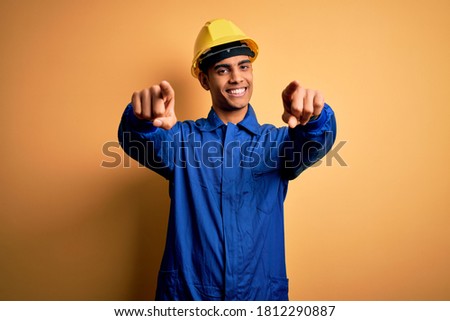 Young handsome african american worker man wearing blue uniform and security helmet pointing to you and the camera with fingers, smiling positive and cheerful