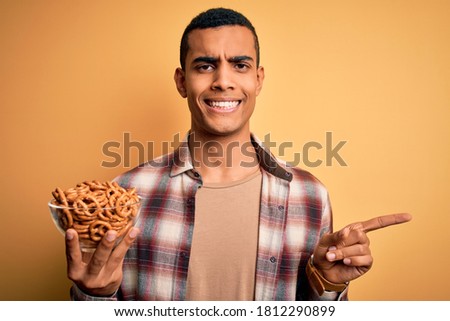 Young handsome african american man holding bowl with german baked pretzels very happy pointing with hand and finger to the side