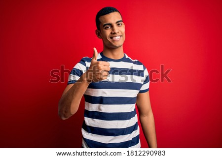 Handsome african american man wearing casual striped t-shirt standing over red background doing happy thumbs up gesture with hand. Approving expression looking at the camera showing success.