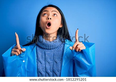 Young beautiful chinese female wearing rain coat standing over isolated blue background amazed and surprised looking up and pointing with fingers and raised arms.