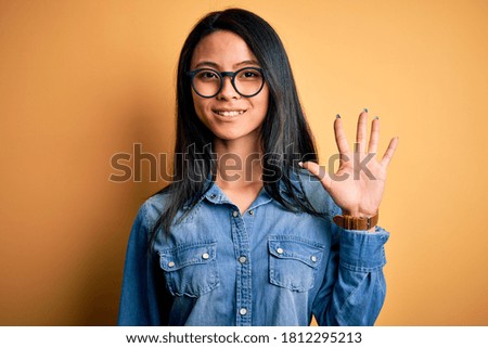 Young beautiful chinese female wearing casual denim shirt over isolated yellow background showing and pointing up with fingers number five while smiling confident and happy.