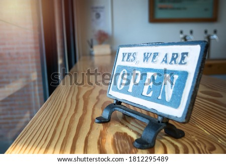 A close-up open sign of a coffee shop lying on a vintage wooden table with blurred background and flare in the top left corner, selective focus.