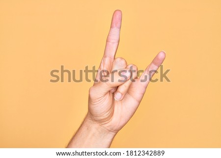 Hand of caucasian young man showing fingers over isolated yellow background gesturing rock and roll symbol, showing obscene horns gesture