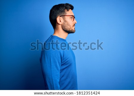 Young handsome man with beard wearing casual sweater and glasses over blue background looking to side, relax profile pose with natural face and confident smile.