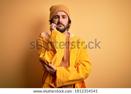 Young handsome man with beard wearing raincoat for rainy day over yellow background thinking looking tired and bored with depression problems with crossed arms.