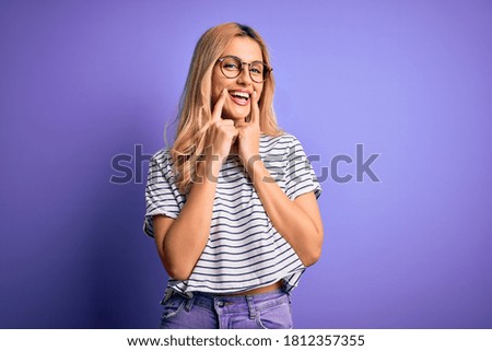 Young beautiful blonde woman wearing striped t-shirt and glasses over purple background Smiling with open mouth, fingers pointing and forcing cheerful smile