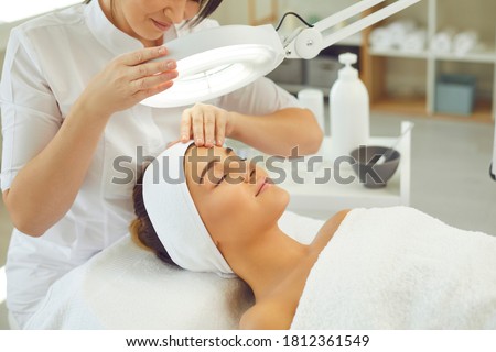 Curing skin problems. Cropped female cosmetologist looking at client's face through magnifying lamp examining her skin. Happy relaxed young woman getting professional facial treatment in spa salon Royalty-Free Stock Photo #1812361549