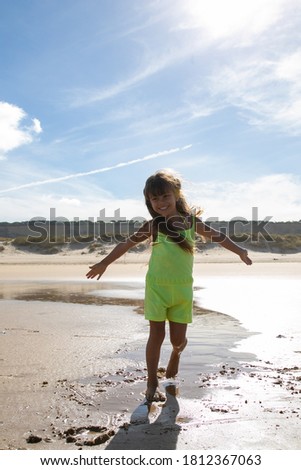 Joyful little girl wearing summer clothes, posing with open arms on wet sand and dancing on beach. Full length. Childhood and leisure outdoors concept