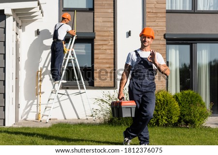Selective focus of handyman with toolbox showing ok at camera while standing on lawn near building