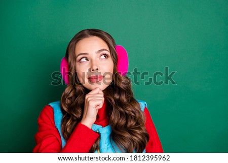 Photo of cute charming young lady smiling finger chin pensive look up empty space thinking ski resort wear pink ear warmers blue vest red pullover isolated green color background