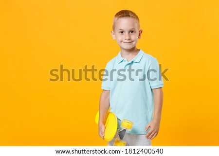 Smiling little fun male fair-haired brown-eyed kid boy 5-6 years old wearing stylish blue turquoise t-shirt polo hold in hands skate board isolated on yellow color background, child studio portrait