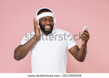 Cheerful young bearded african american fitness sports man 20s in white headband t-shirt listening music with headphones using mobile phone isolated on pastel pink color background studio portrait